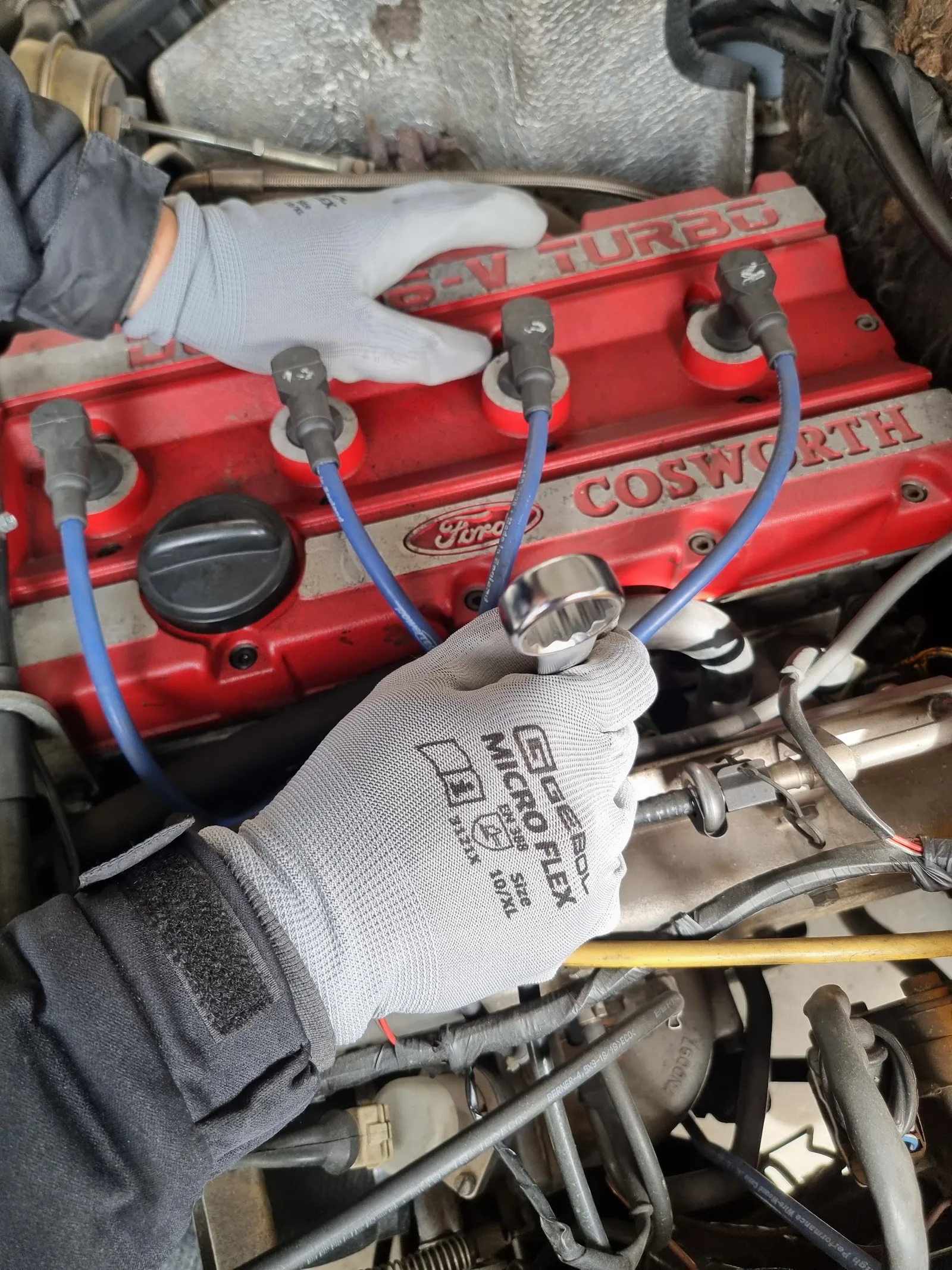 Zwei Hände in Arbeitshandschuhen am roten Ventildeckel eines Ford-Cosworth-16V-Turbo-Motors, blaue Zündkabel und ein Steckschlüsseleinsatz