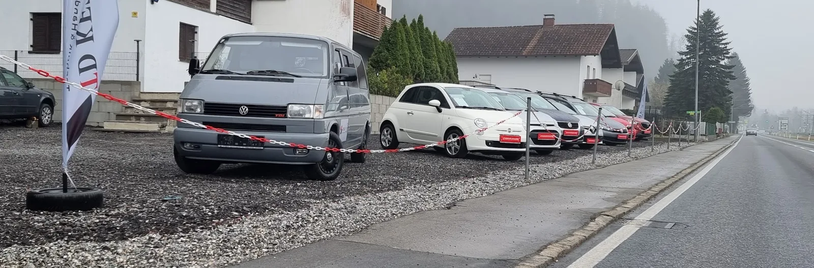 Der Verkaufsplatz an der Straße mit einem VW-Bus, einem Fiat 500 und weiteren Fahrzeugen hinter einer rot-weißen Kette, daneben die Fahne des Betriebs