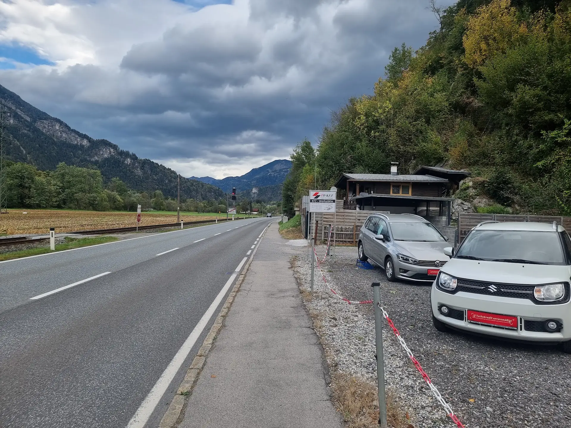 Der Betrieb von der Landstraße aus: Firmenschild mit der Aufschrift TD KFZ, Fahrzeuge auf dem Platz und die Berge des Montafon